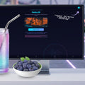 A computer monitor on a desk displaying a software interface titled 'Pantry OS' with a tropical sunset graphic and promotional text. In front of the monitor is a white bowl of blueberries and a glass with a metal straw and pastel drink, set against a blurred modern interior background.